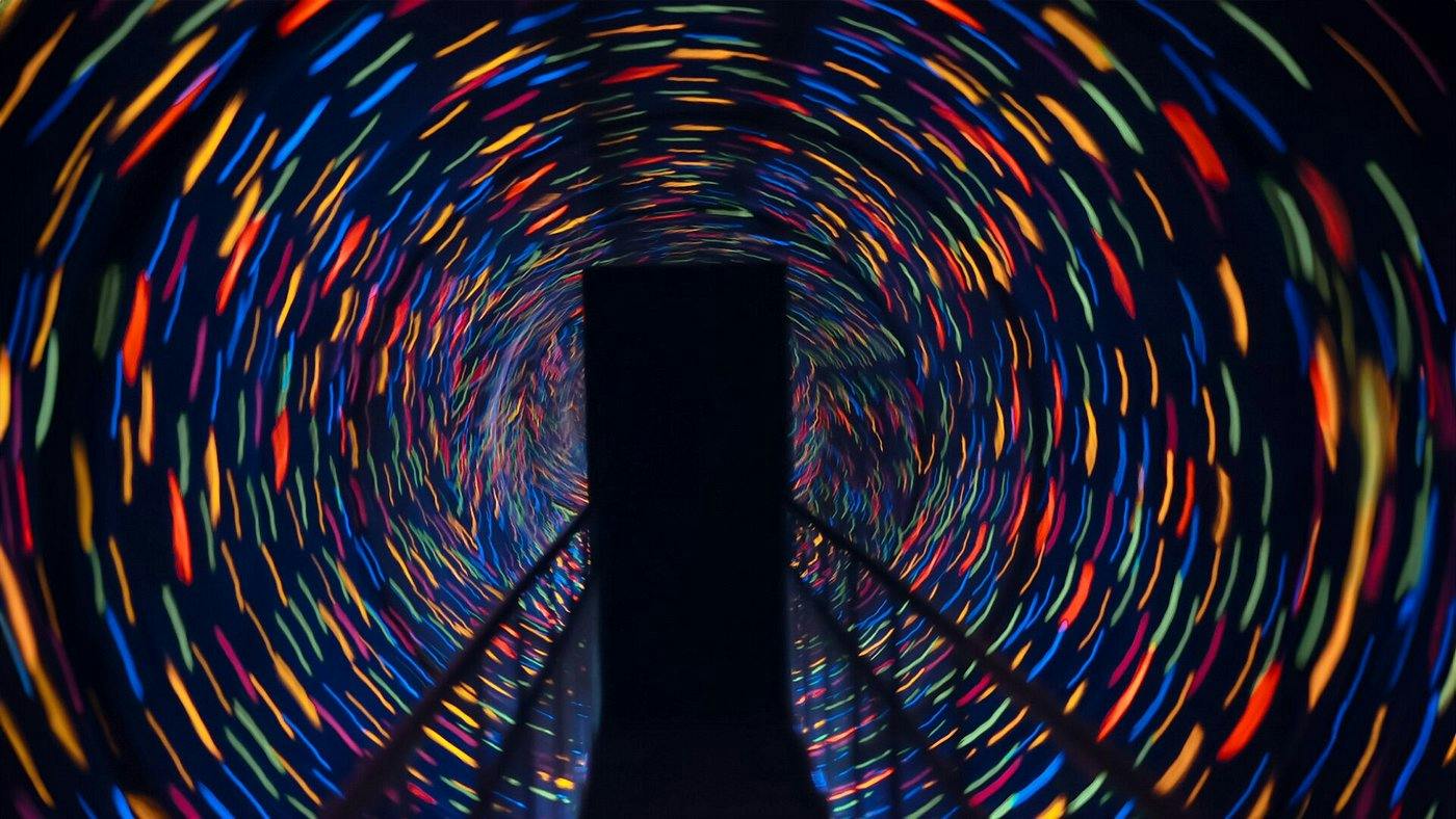 A tunnel with swirling multicolored lights creating a spiraling effect, viewed from a walkway with railings.