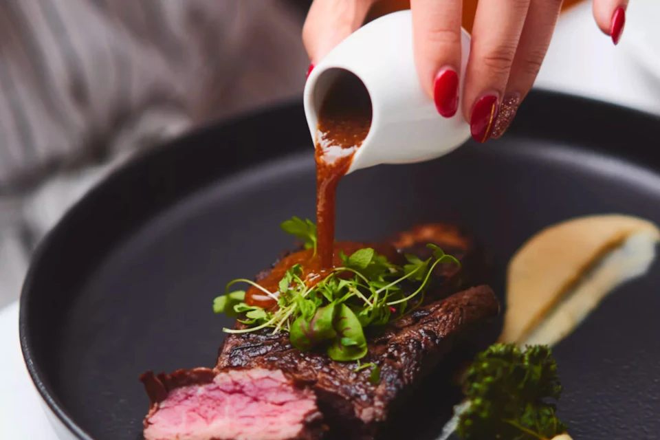 A hand with red nail polish pours sauce from a white jug onto steak garnished with greens on a black plate.