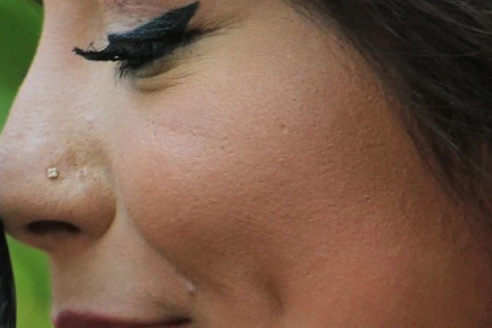 Woman with winged eyeliner and a nose piercing smiling closely at a dark gray parrot with red cheek patches.