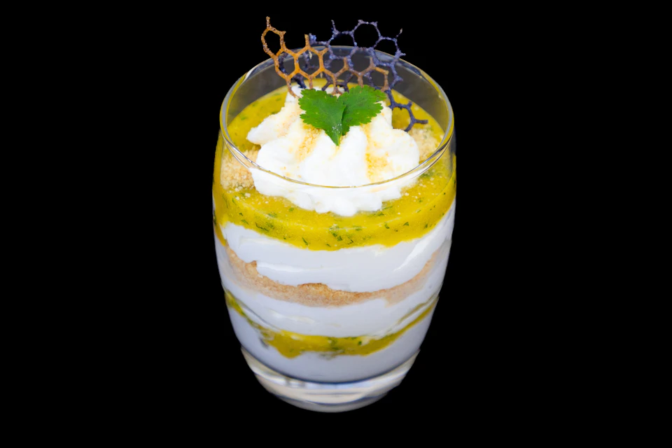 A layered dessert in a glass with whipped cream, yellow sauce, crumbles, and decorative honeycomb garnish on top.