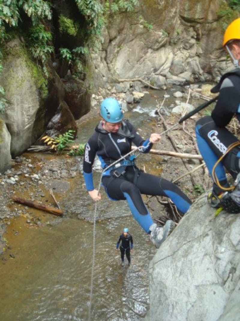 Люди в гидрокостюмах и шлемах спускаются по скалистой скале над ручьем в лесистом каньоне.