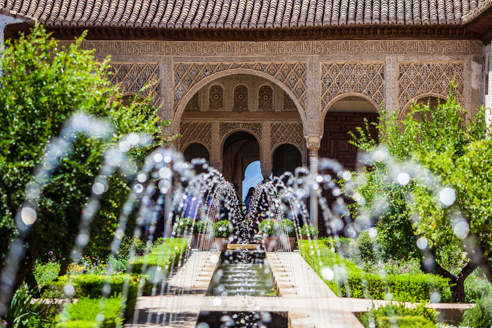 Ornate stone archways behind a central fountain with water jets, surrounded by lush greenery and manicured bushes.