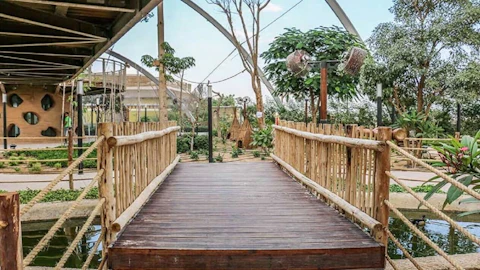 A wooden bridge with rope railings leads to a lush garden area, featuring various plants, trees, and a structure with circular windows.