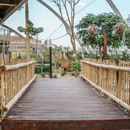A wooden bridge with rope railings leads to a lush garden area, featuring various plants, trees, and a structure with circular windows.