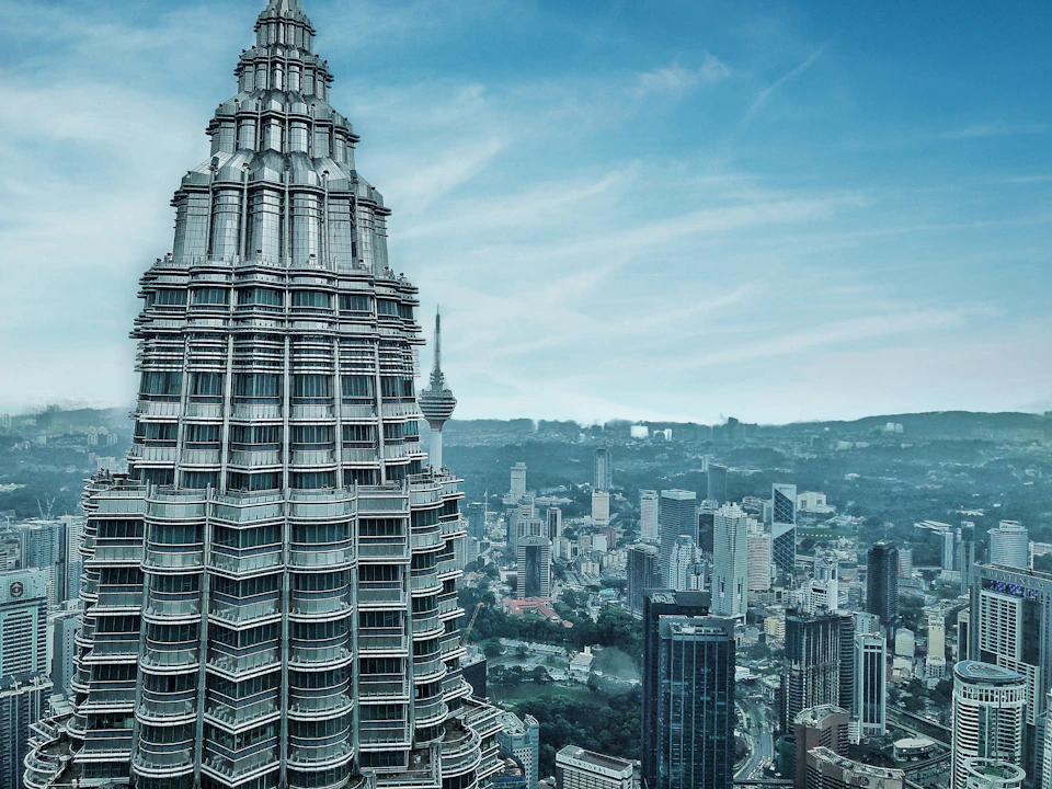 A view of the Petronas Towers in Kuala Lumpur, with the cityscape and Kuala Lumpur Tower visible under a clear blue sky.
