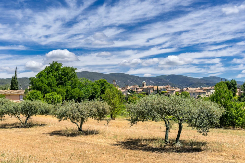 Countryside landscape with olive trees, a village in the distance, and mountains under a partly cloudy blue sky.