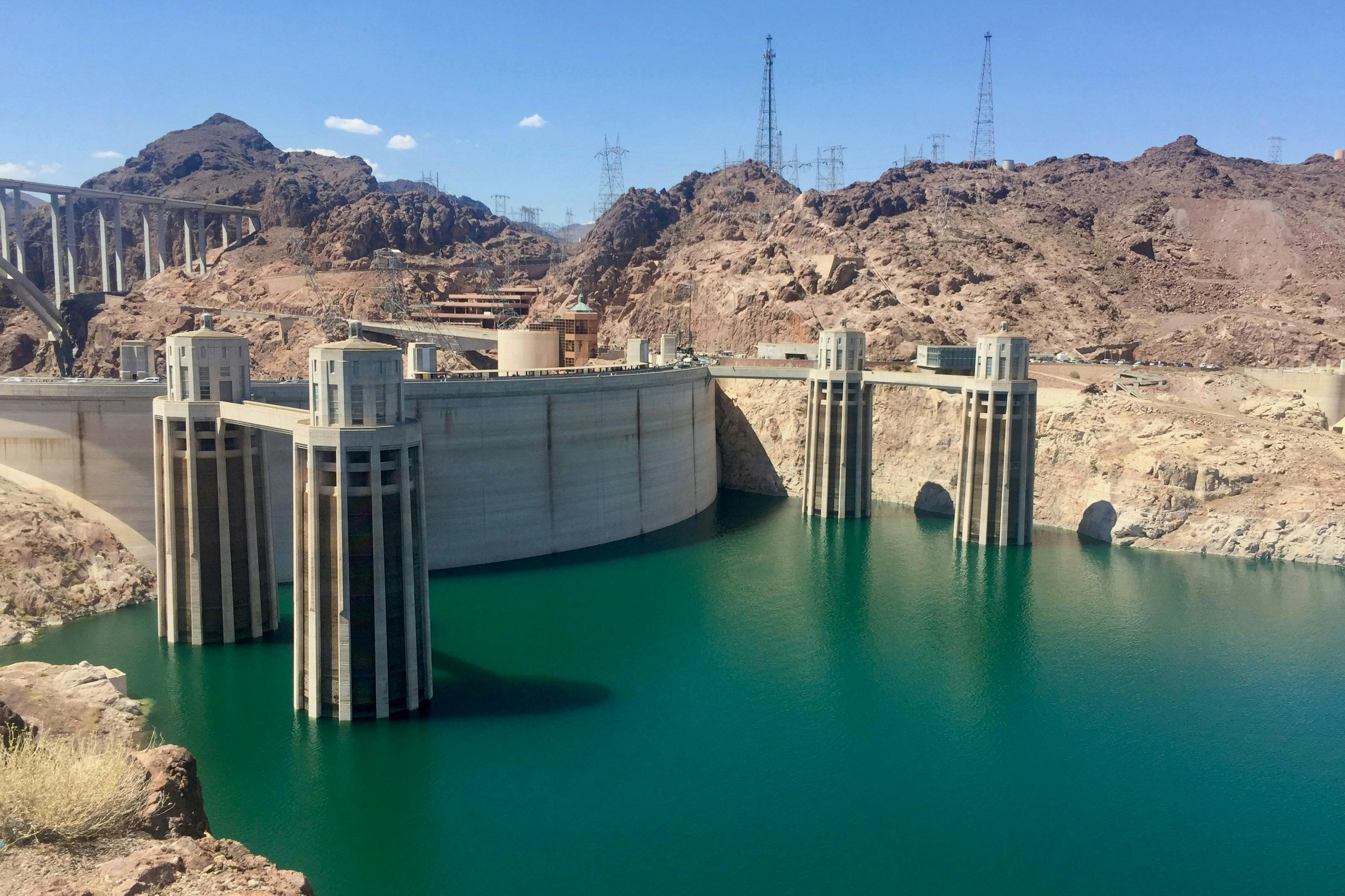 A large concrete dam spanning a reservoir with turquoise water, surrounded by rocky desert terrain under a clear sky.