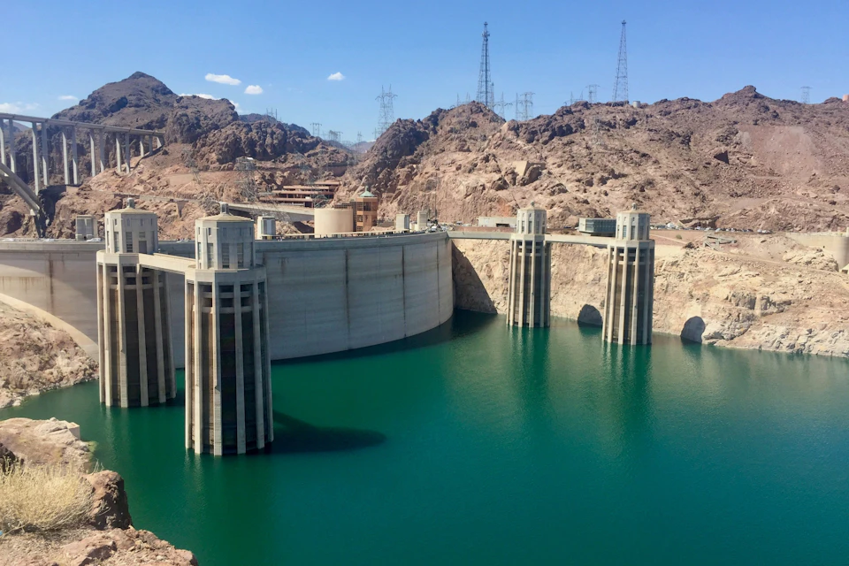 A large concrete dam spanning a reservoir with turquoise water, surrounded by rocky desert terrain under a clear sky.