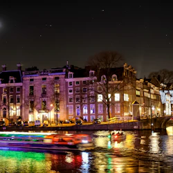Upplyst kanal i Amsterdam på natten, med en förbipasserande båt, ljusdekorerad bro, måne på himlen och byggnader i bakgrunden.