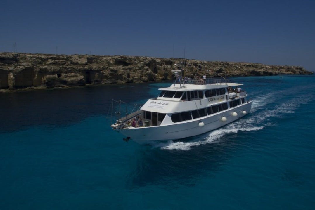 Ein weißes Touristenboot, das unter klarem Himmel durch blaues Wasser mit felsiger Küste im Hintergrund fährt.