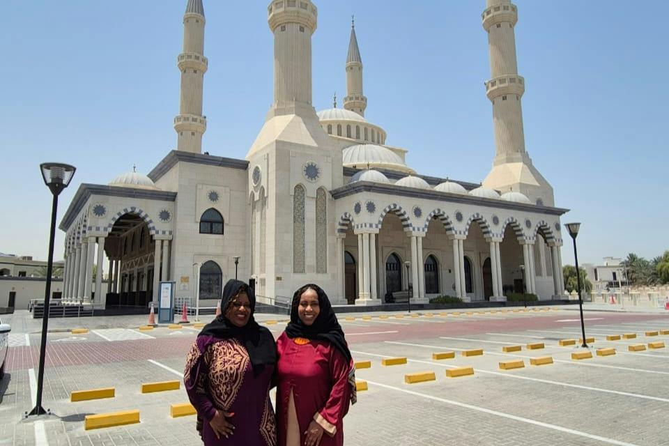 Two women in colorful attire standing in front of a large mosque with tall minarets on a sunny day.