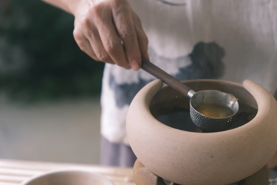A person holding a ladle over a ceramic bowl filled with liquid, with a blurred background.