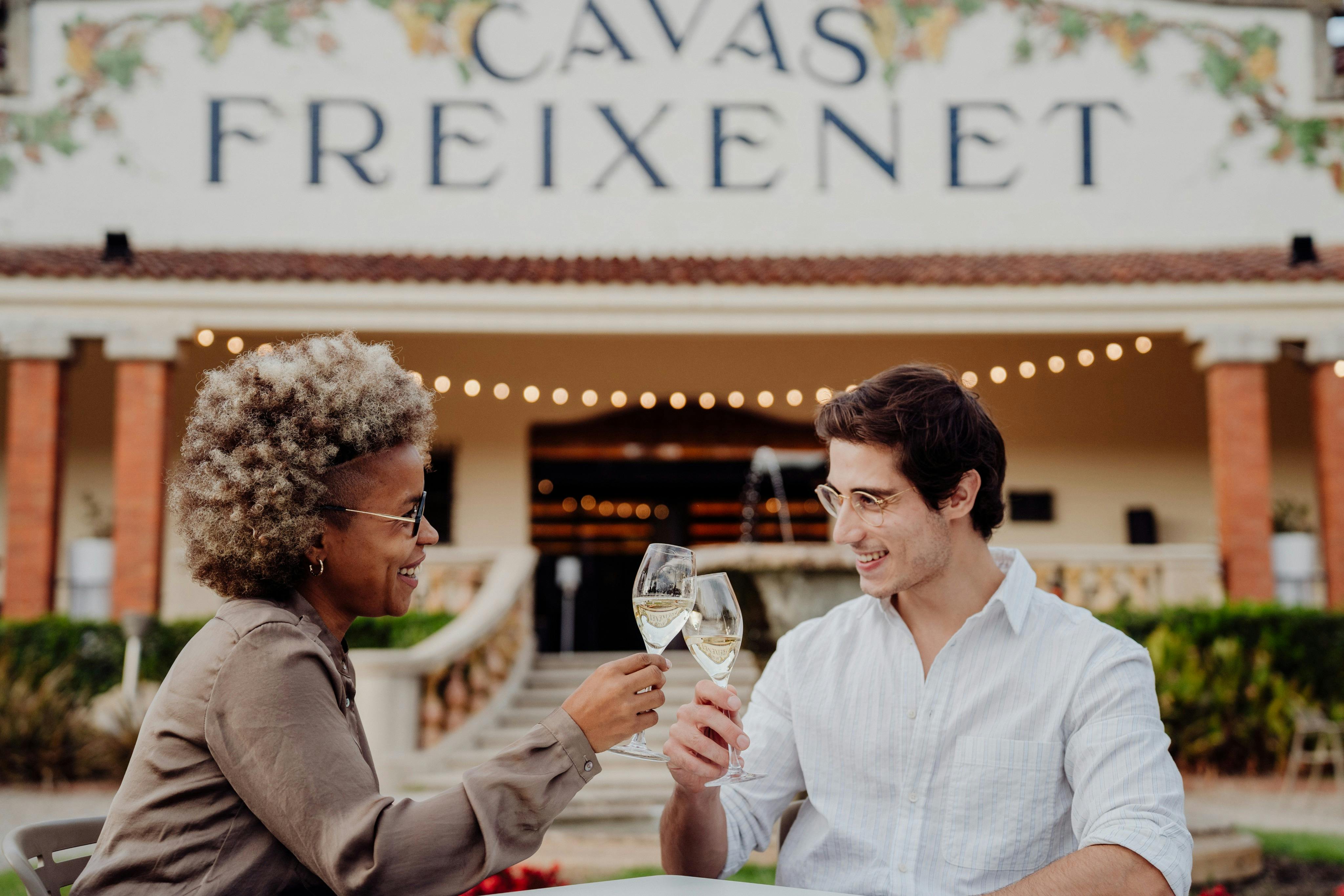Un hombre con camisa blanca brinda con alguien fuera de cuadro, sosteniendo un vaso de vino blanco delante de un edificio con el rótulo "Cavas Freixenet".