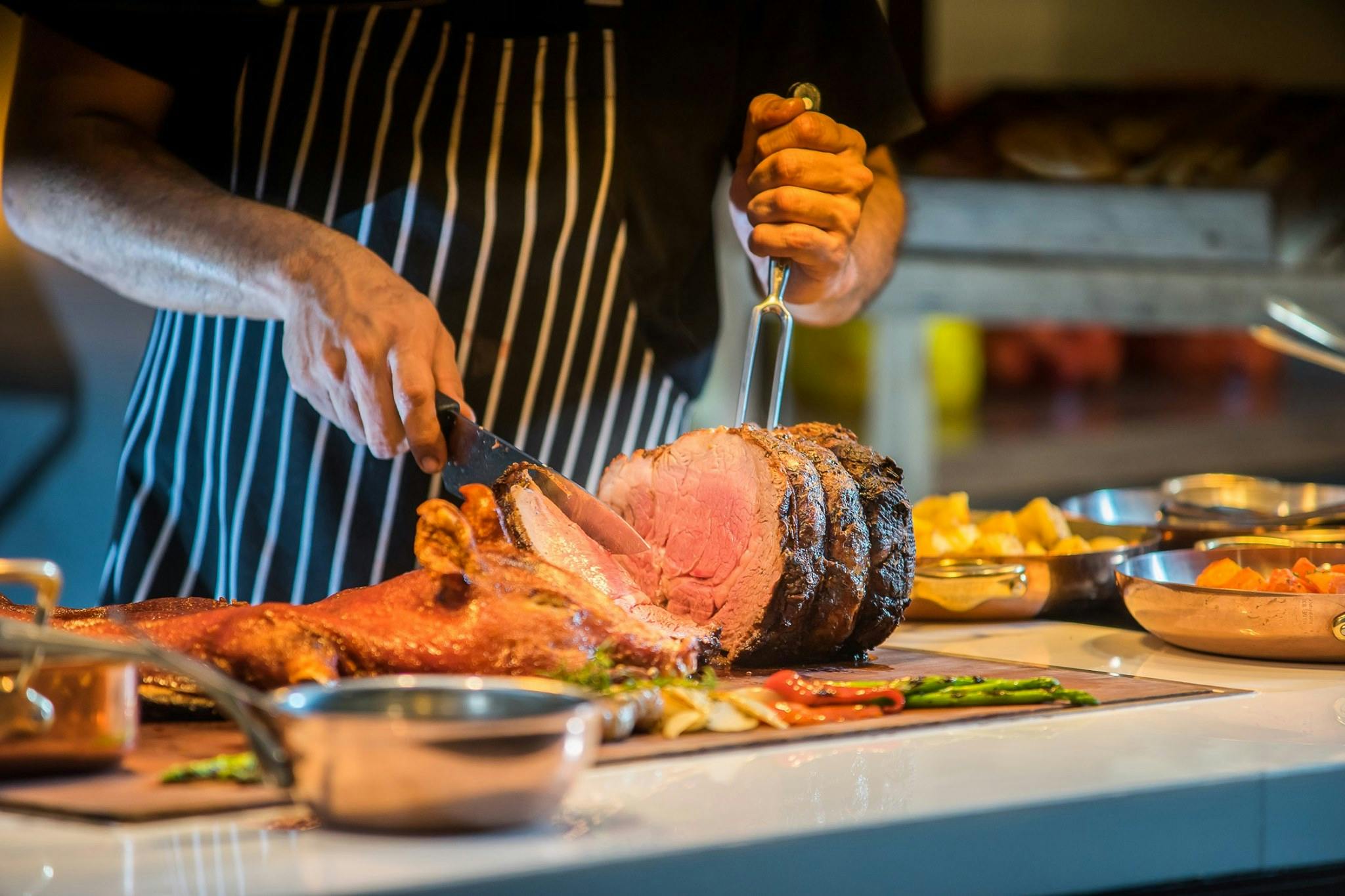 A chef carving a roasted meat joint on a buffet table with vegetables and side dishes displayed around it.