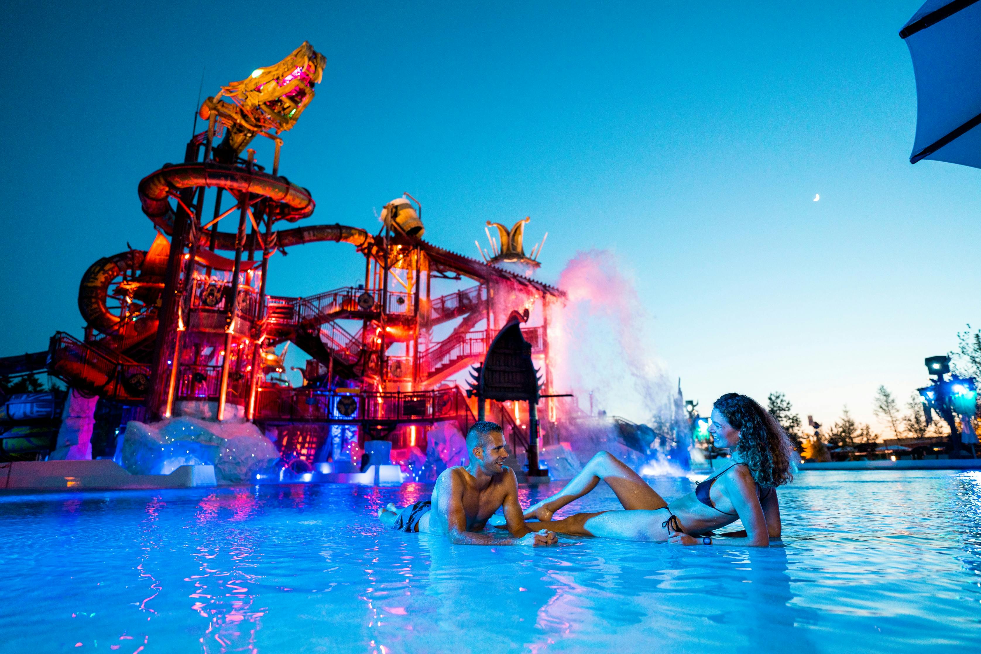 Dos personas en bañador se relajan en una piscina de un parque acuático iluminada y llena de color al anochecer, con unos imponentes toboganes al fondo.
