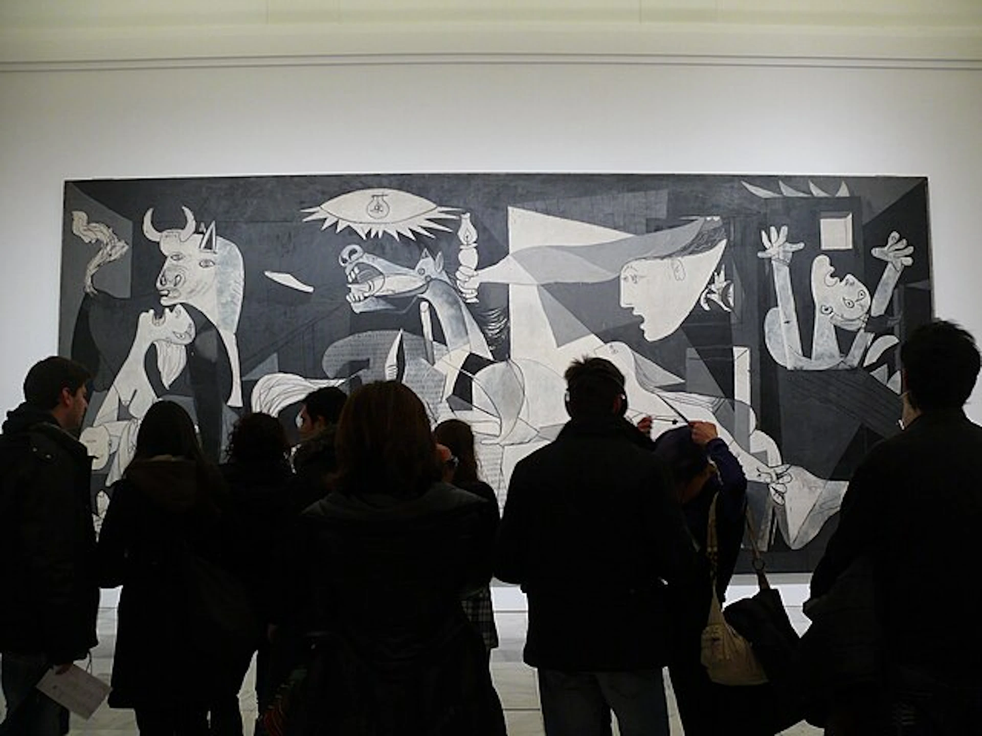 A group of people stand in front of Pablo Picasso's large, monochromatic painting "Guernica" in a well-lit gallery.