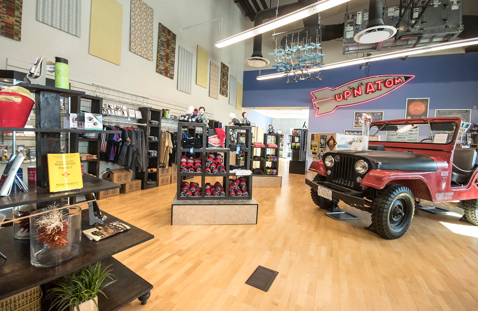 Interior of a shop with clothing racks, shelves of merchandise, and a red jeep display under a pink neon "UPNATO" sign.