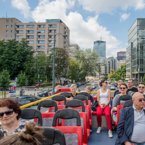 Människor som sitter på det övre däcket på en dubbeldäckad buss, med en stadsbild av byggnader och träd i bakgrunden.