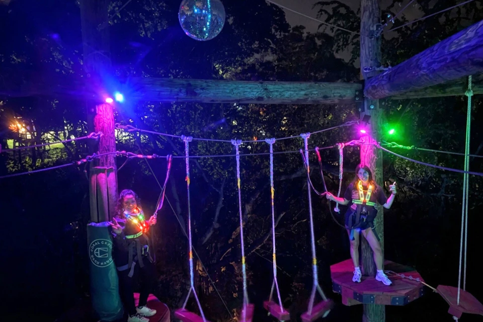 Two people wearing harnesses and neon lights standing on a rope course platform at night, surrounded by trees.