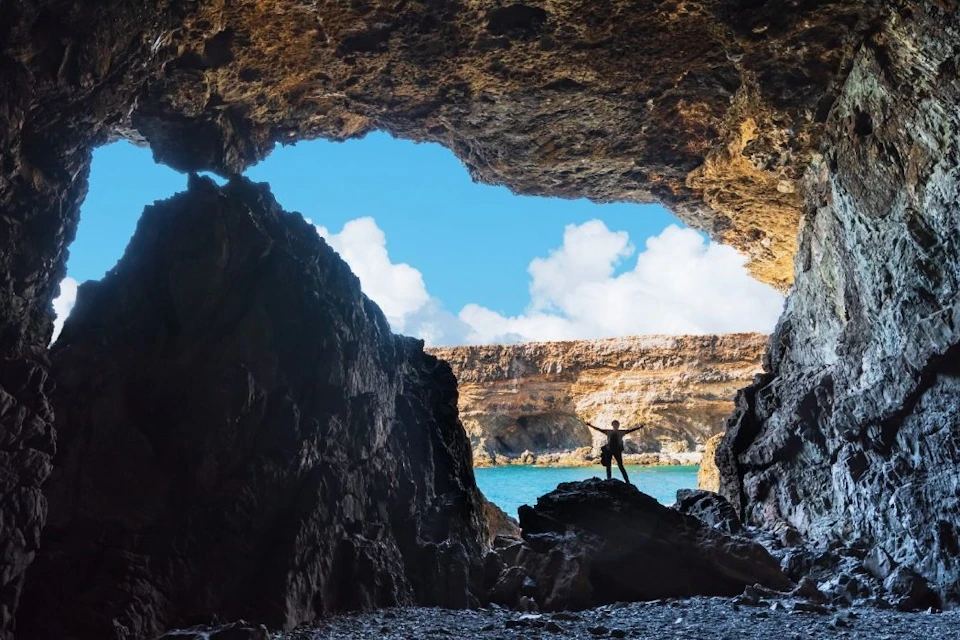 A person stands on rocks at the entrance of a cave overlooking a blue sky with clouds and a distant cliff.