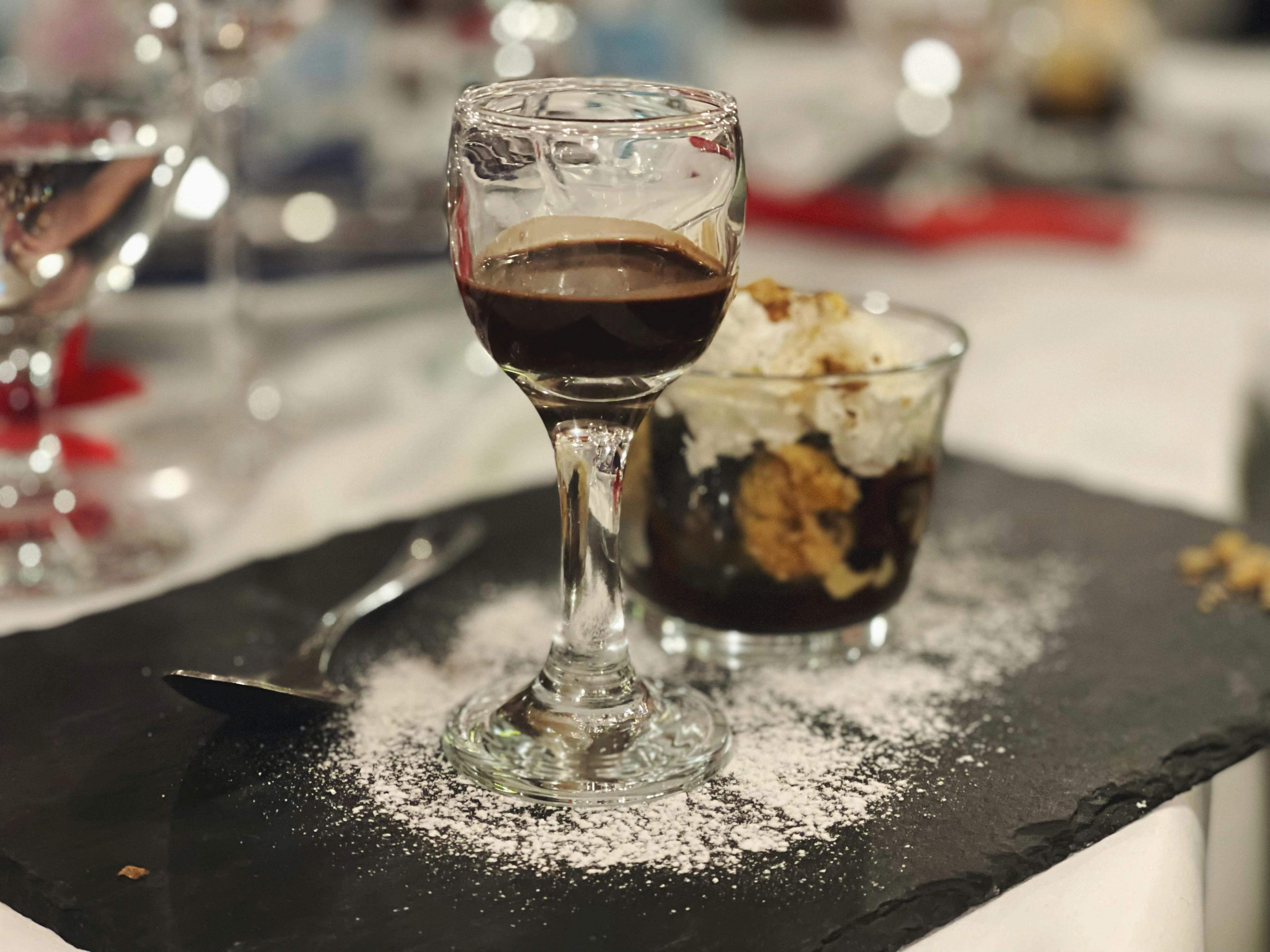 Ένα ποτήρι σκούρο υγρό και ένα επιδόρπιο σε ένα γυάλινο πιάτο, τοποθετημένα σε μια πλάκα από σχιστόλιθο πασπαλισμένα με ζάχαρη άχνη. Κουτάλι στο πλάι.