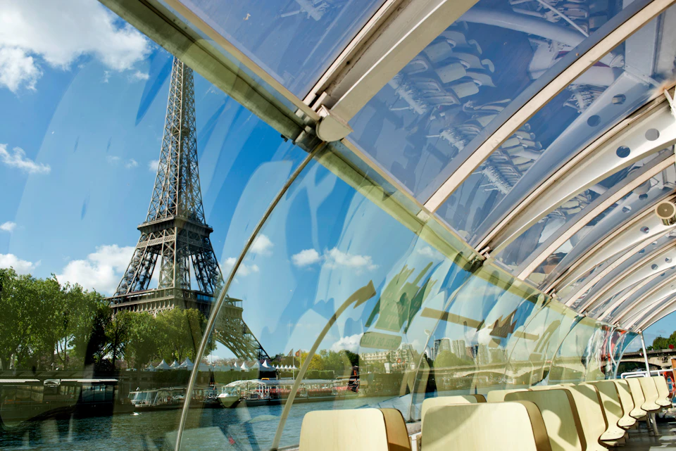 View of the Eiffel Tower from inside a glass-roofed boat on the Seine River, with empty seats and reflections visible.