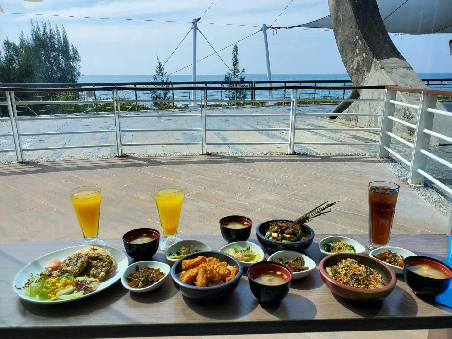 A table set with various dishes, drinks, and side dishes, overlooking a seaside view with railings and a clear sky.