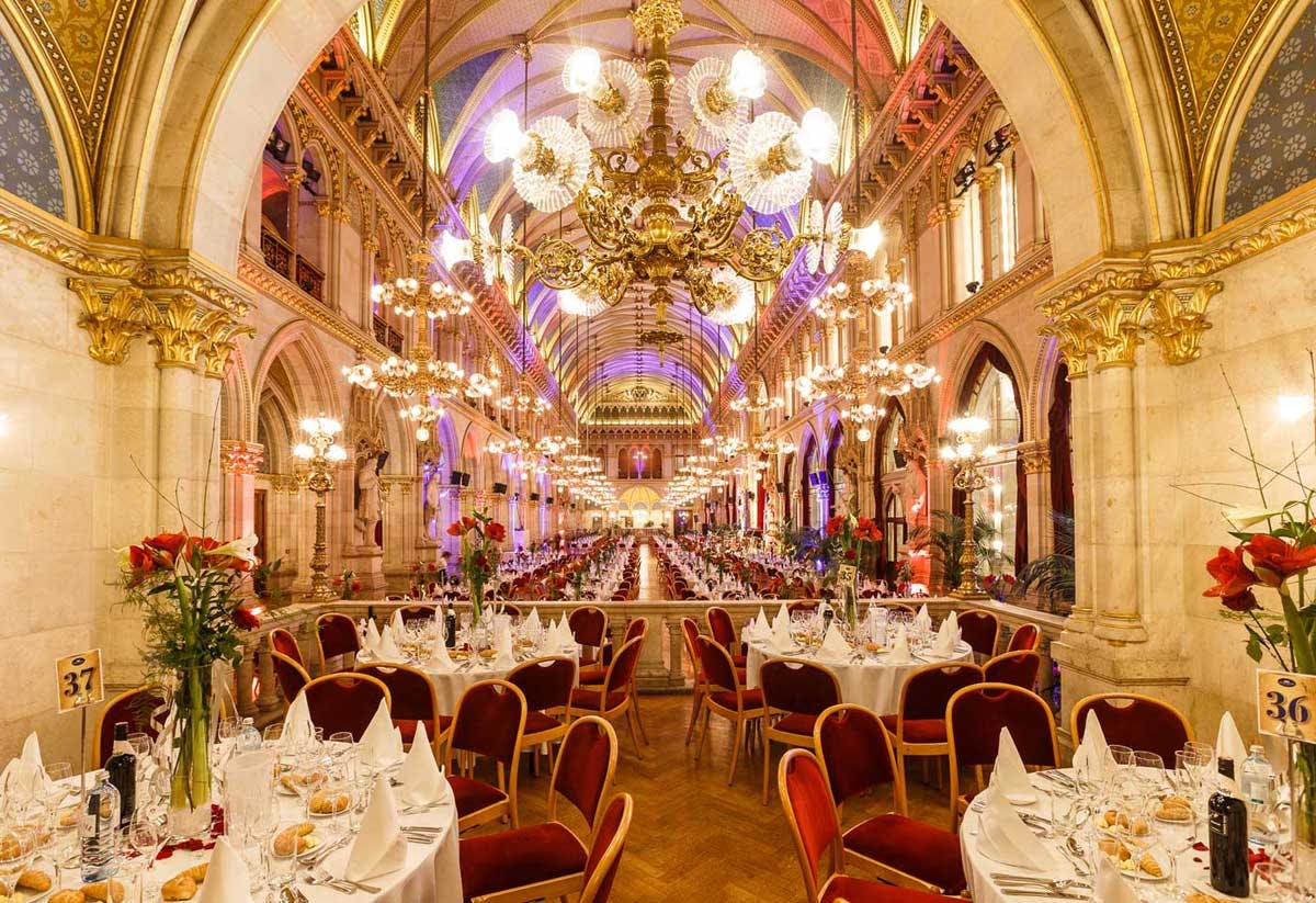 Πολυτελώς διακοσμημένη αίθουσα δεξιώσεων με κόκκινες βελούδινες καρέκλες, επίσημα τραπέζια, πολυελαίους και τοξωτές οροφές.