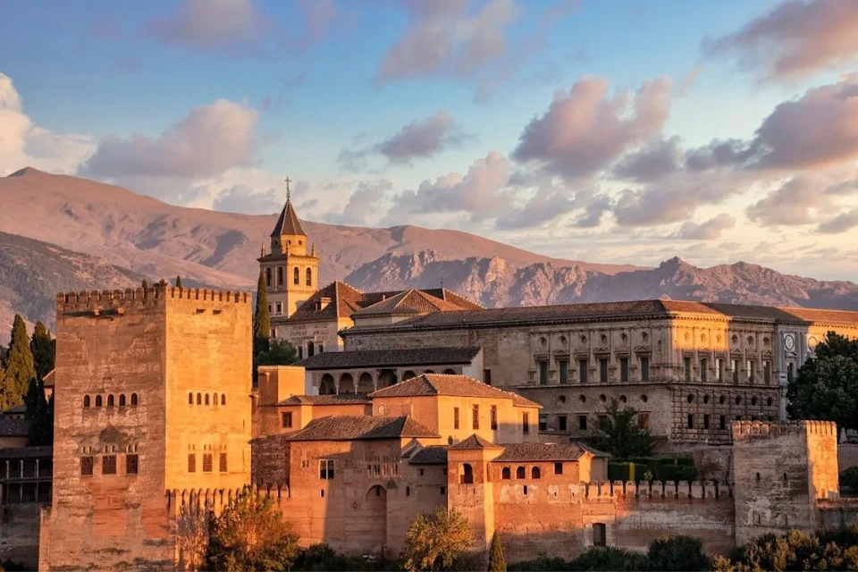 Historic stone buildings with tiled roofs and a tower, set against a mountainous backdrop under a partly cloudy sky.