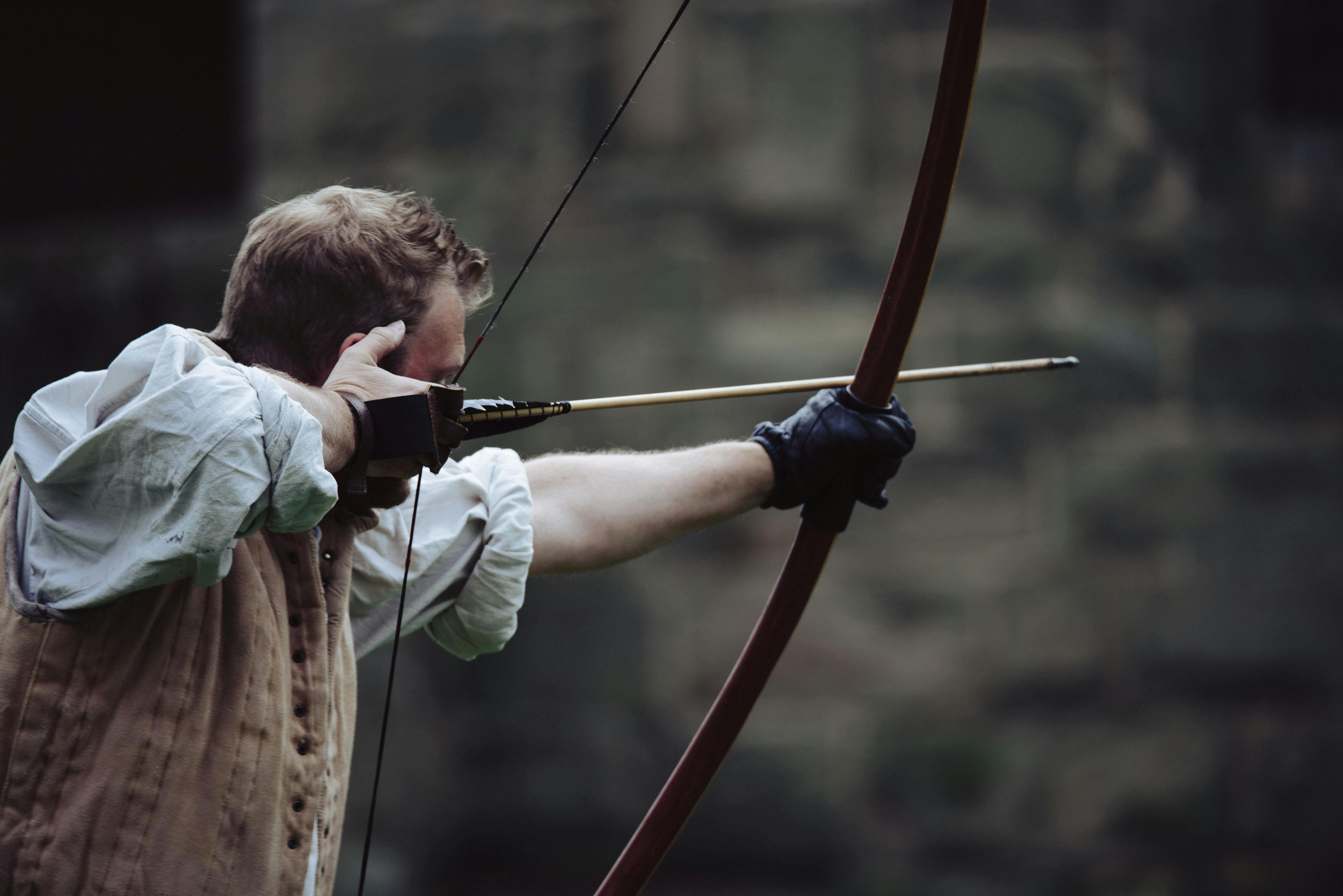 A person in historical attire is drawing a bow with an arrow, wearing a glove on one hand. Stone wall in the background.