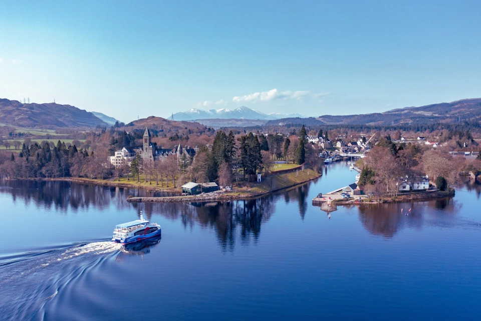 A scenic lakeside town with a ferry on the water, surrounded by trees and mountains under a clear blue sky.