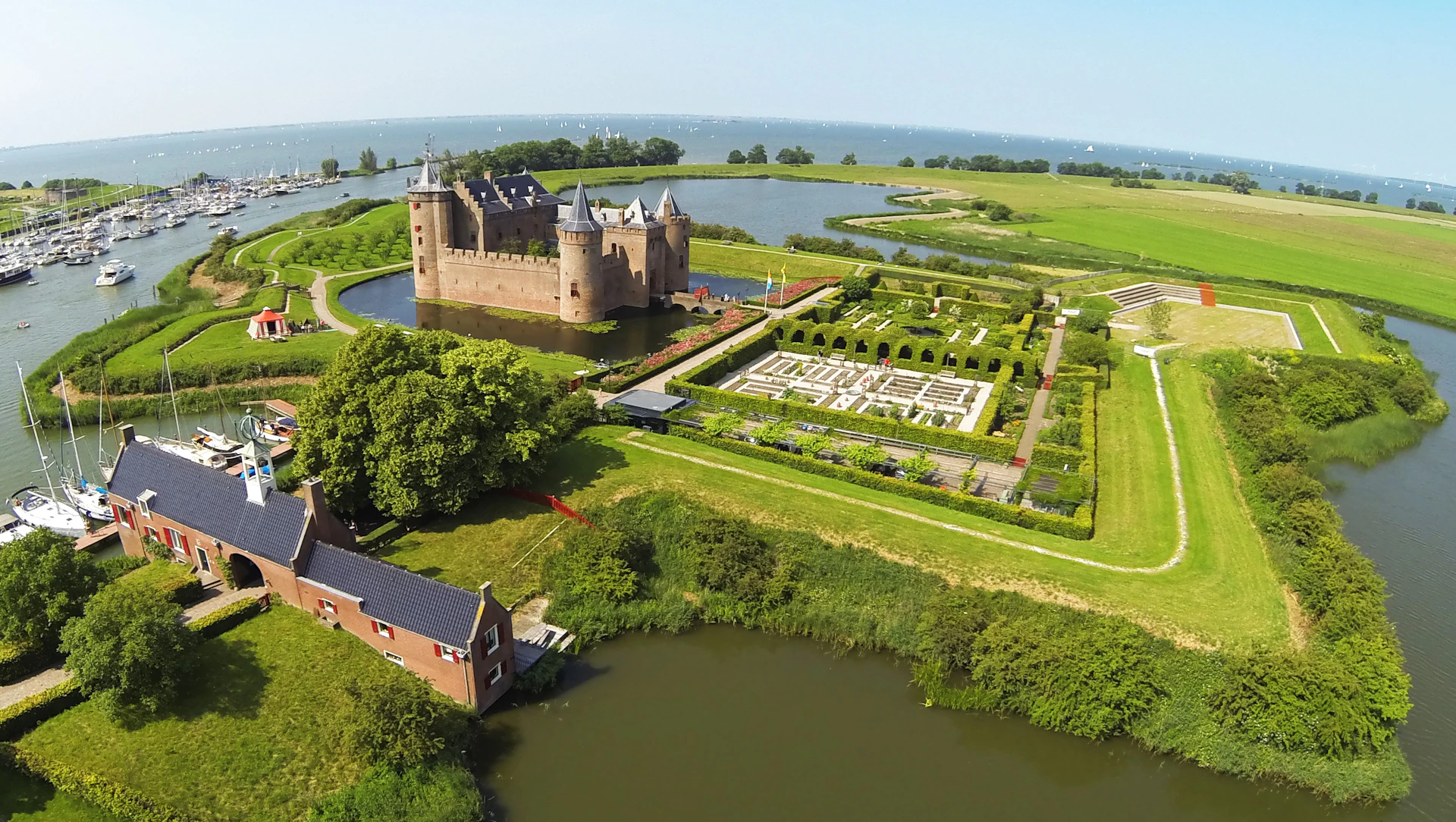 Aerial view of a castle with a moat, adjoining gardens, a body of water in the background, and a smaller building nearby.