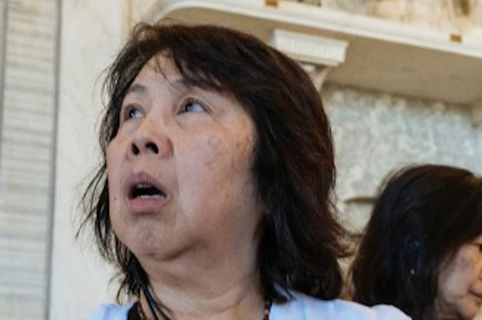 A group of people in a room with ornate walls and artwork, one woman holding an orange bag and looking upwards.