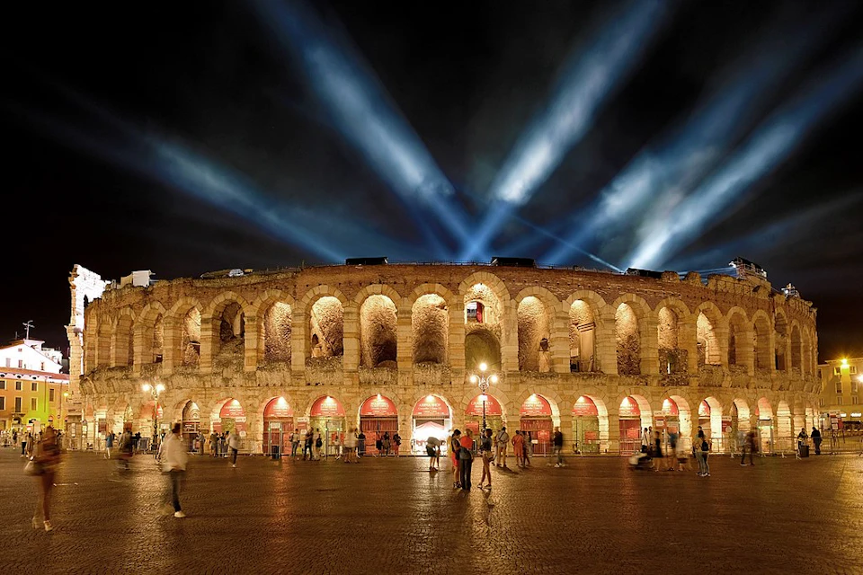 Ancient amphitheater illuminated at night with spotlights shining upwards, people walking in the foreground.