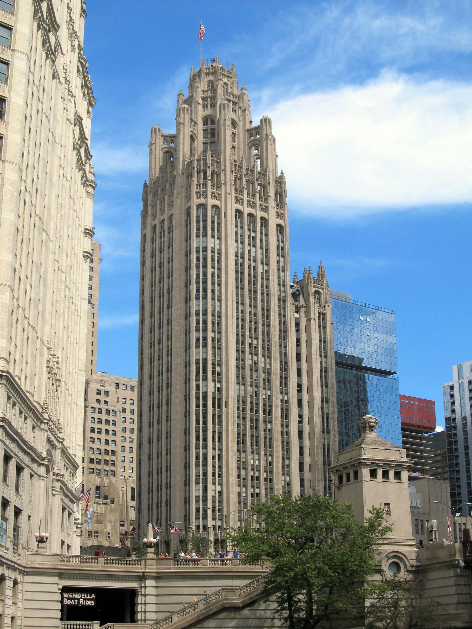 Tall, ornate building with American flag on top, surrounded by other buildings, under a blue sky with some clouds.