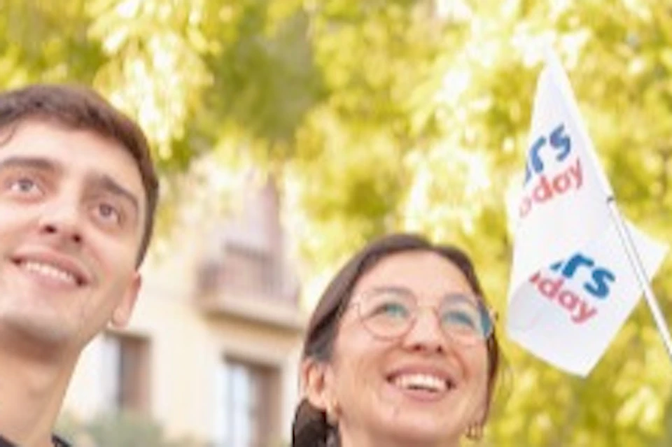 Four people outdoors, smiling and looking upward. One holds a small flag that reads "RS Today." Trees are in the background.
