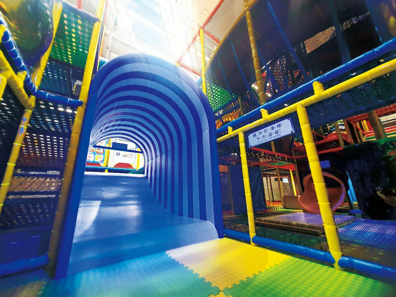 Indoor children's play area with a blue tunnel slide, colorful soft flooring, and climbing structures.