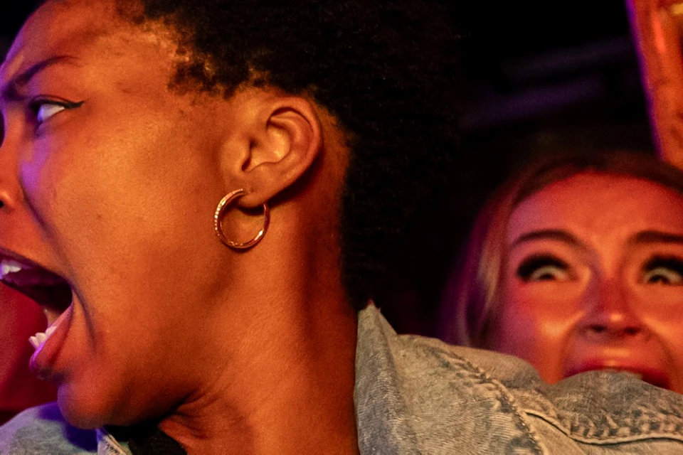 Three people in various expressions of surprise or fear, with scaffolding and dim lighting in the background.