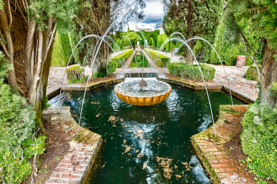 A symmetrical garden fountain with arched water streams, surrounded by brick paths and lush greenery, under a cloudy sky.