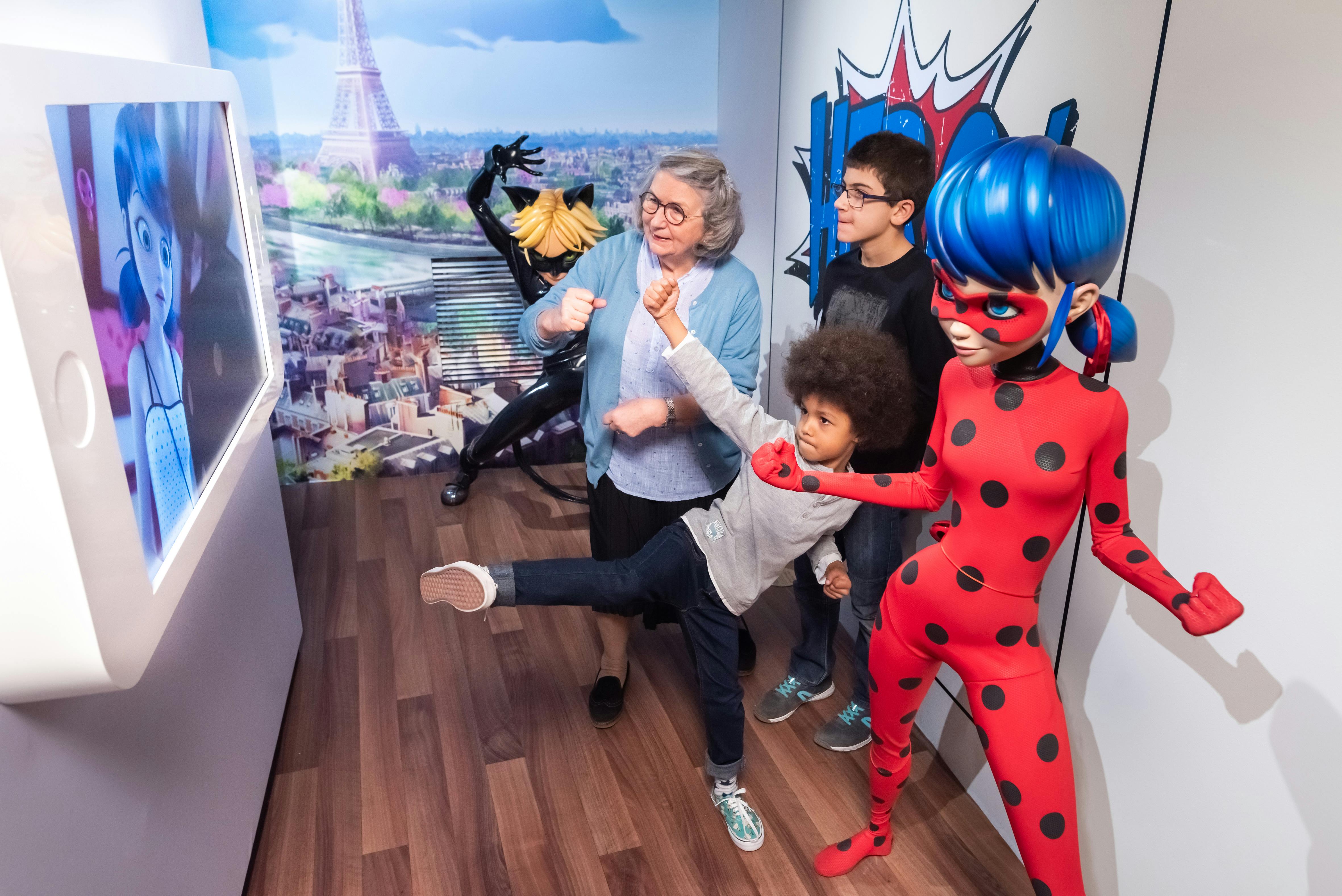 Een vrouw, jongen en kind poseren met de gekostumeerde personages Ladybug en Cat Noir, die allemaal binnenshuis speelse superhelden poses aannemen.