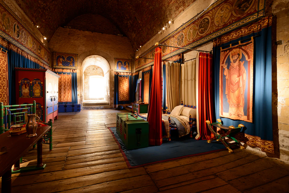 A historical room with wooden floors, decorated with colorful drapes, tapestries, a canopy bed, and medieval-style furnishings.