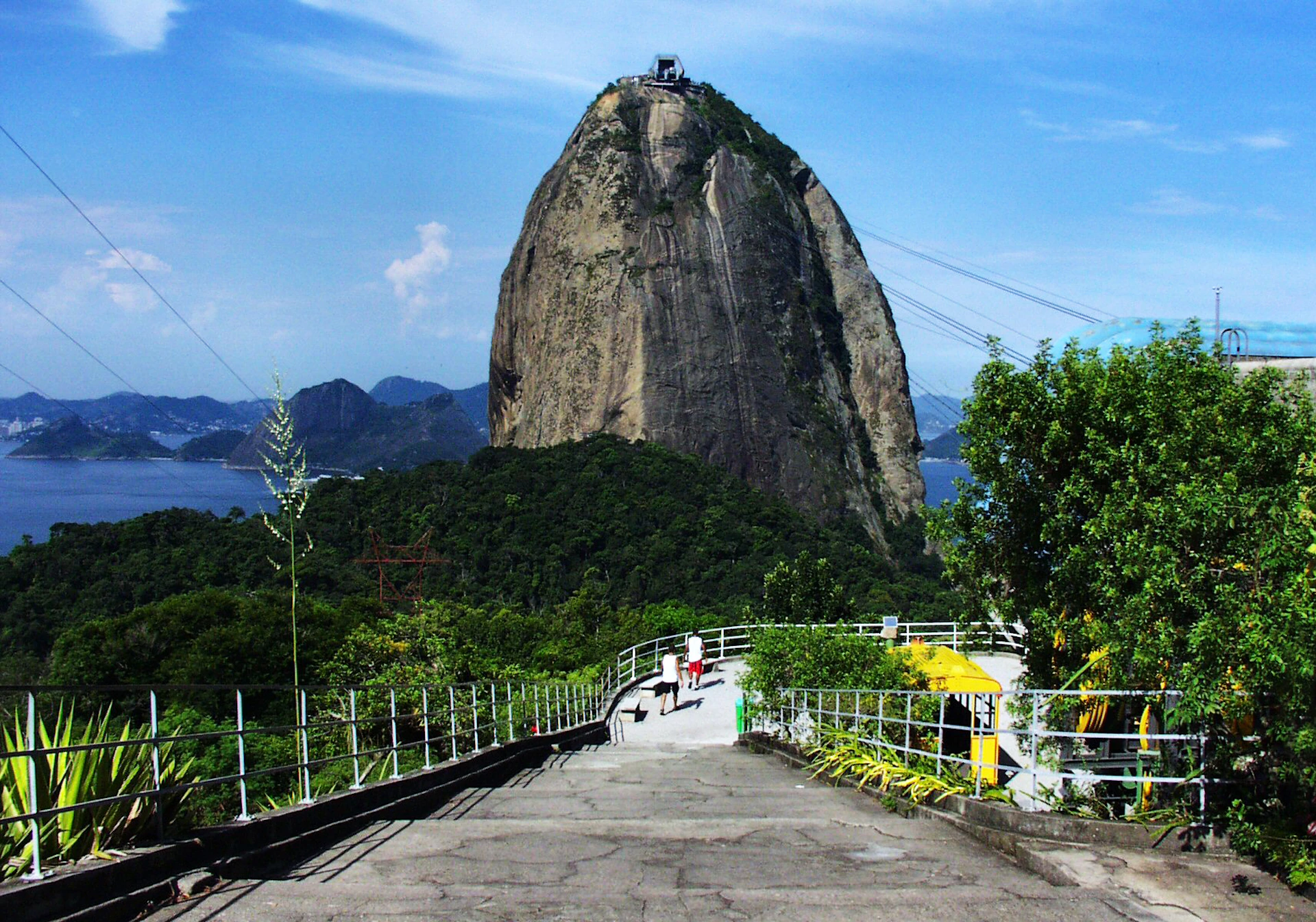 A steep rock formation surrounded by greenery under a blue sky with a paved pathway and railing in the foreground.