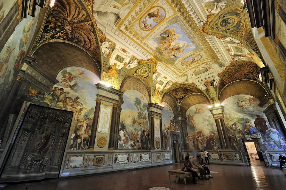 A grand hall with ornate frescoes, gilded details, and a painted ceiling; several people sit and stand, admiring the decor.