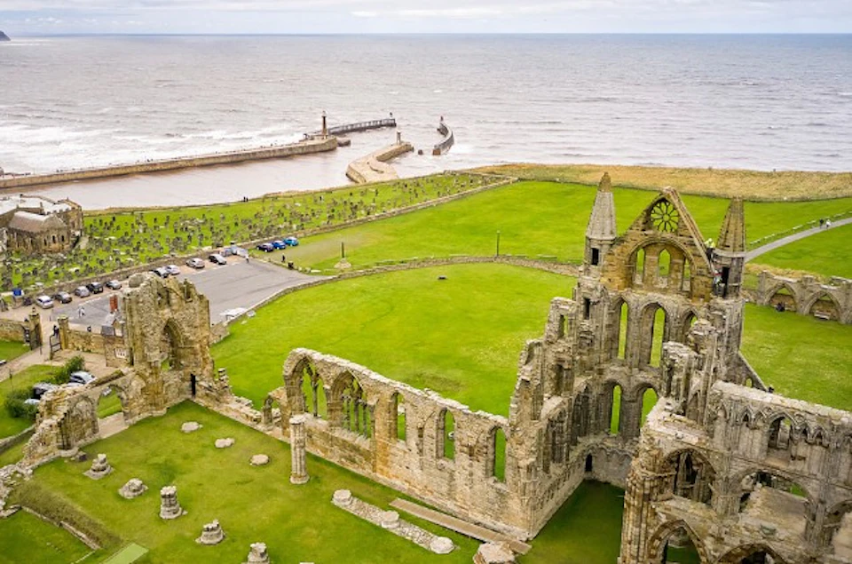 Aerial view of a historic stone ruin on green grass near the sea, with a pier and lighthouse in the background.