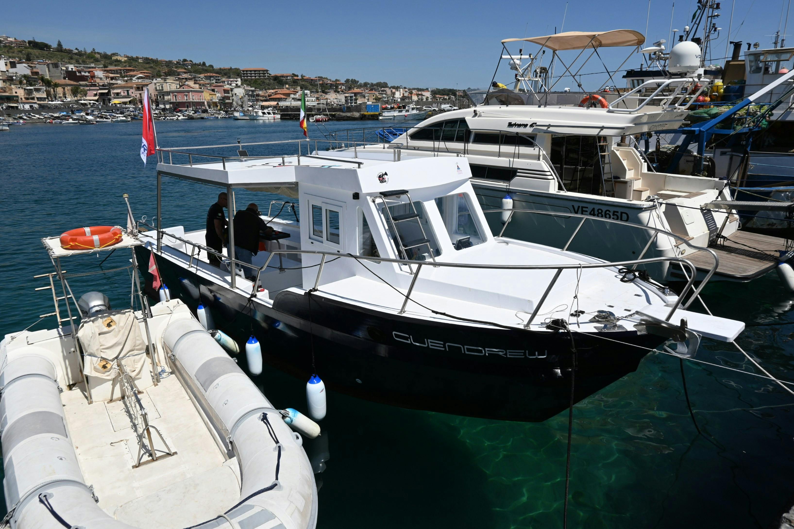 Un pequeño barco blanco llamado "GUENDREW" atracado en un puerto deportivo, con gente a bordo y otros barcos cerca. Al fondo se ven edificios de la ciudad.