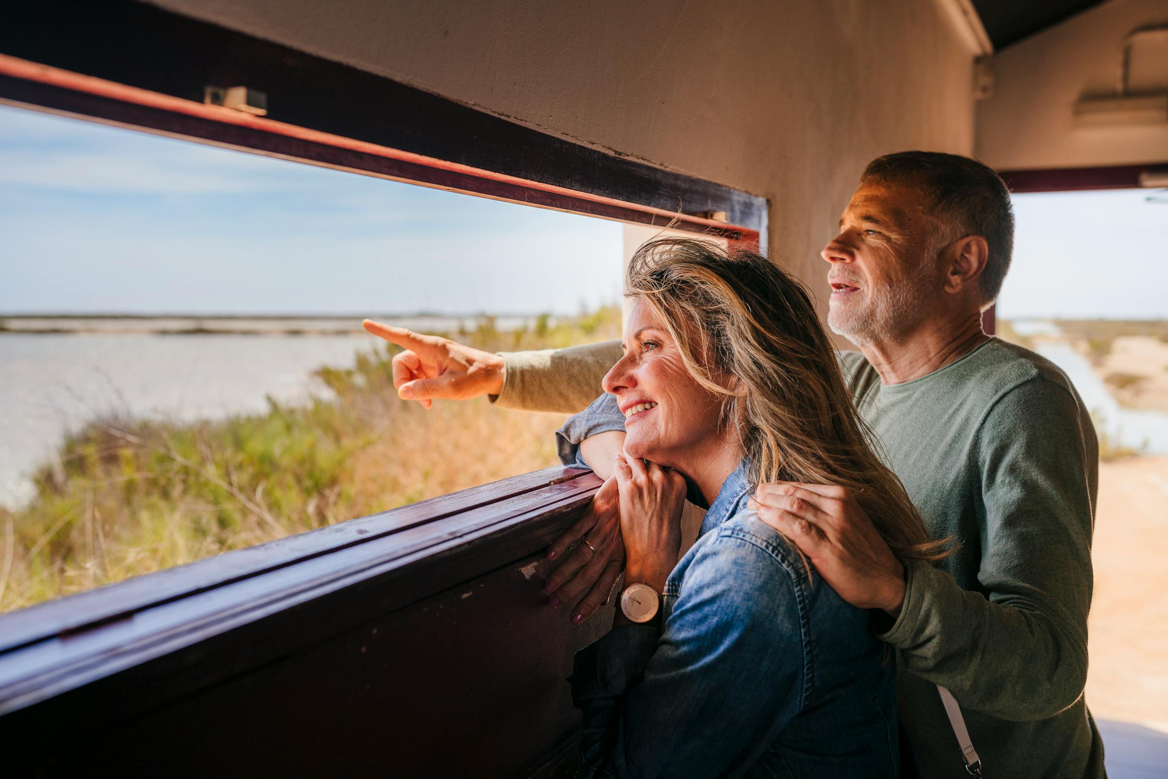 Usmívající se pár se dívá z okna a žena ukazuje na něco v dálce.