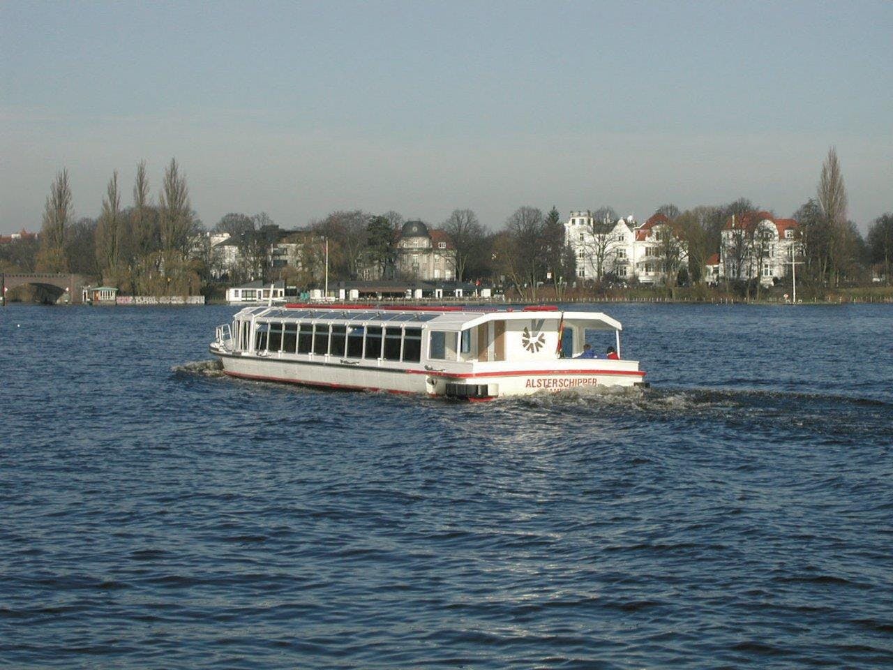 Una barca turistica bianca chiamata "ALSTERSCHIPPER" naviga su uno specchio d'acqua calmo con una riva alberata ed edifici sullo sfondo.