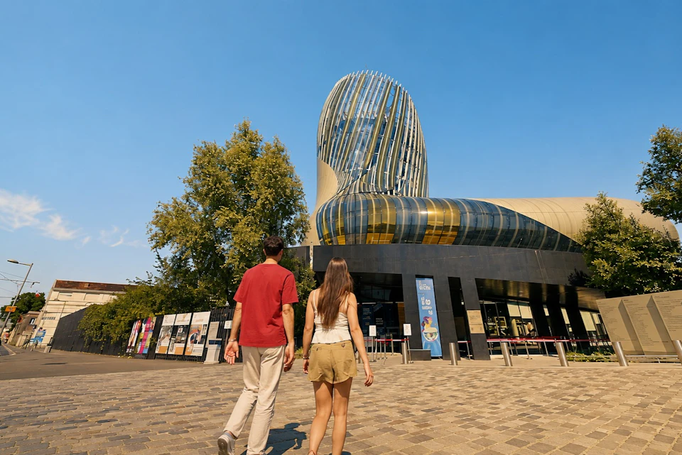 Two people walking toward a modern building with a unique curved design and glass panels under a clear blue sky.