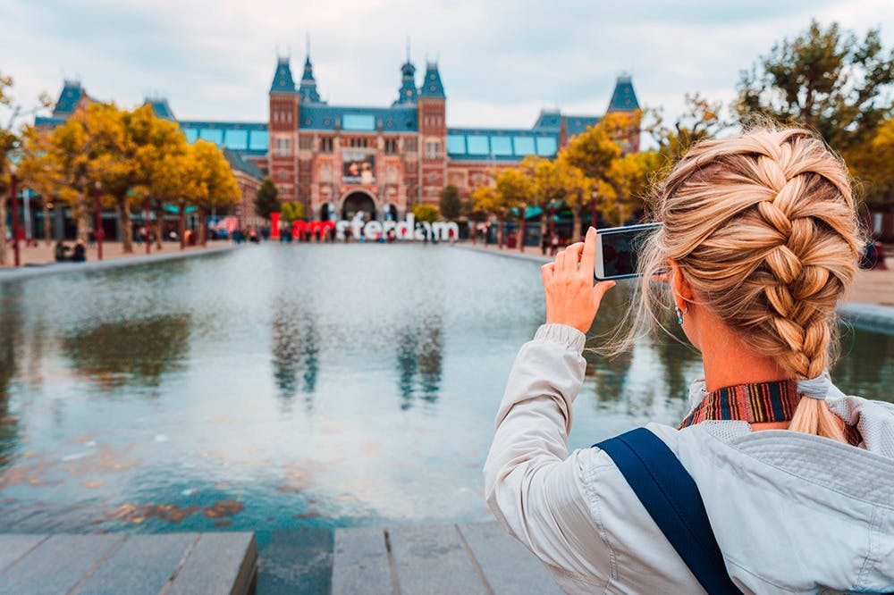 一个人正在拍摄「I amsterdam」标志和一座大型建筑物的照片，该建筑物位于反光泳池对面，两侧有树木。