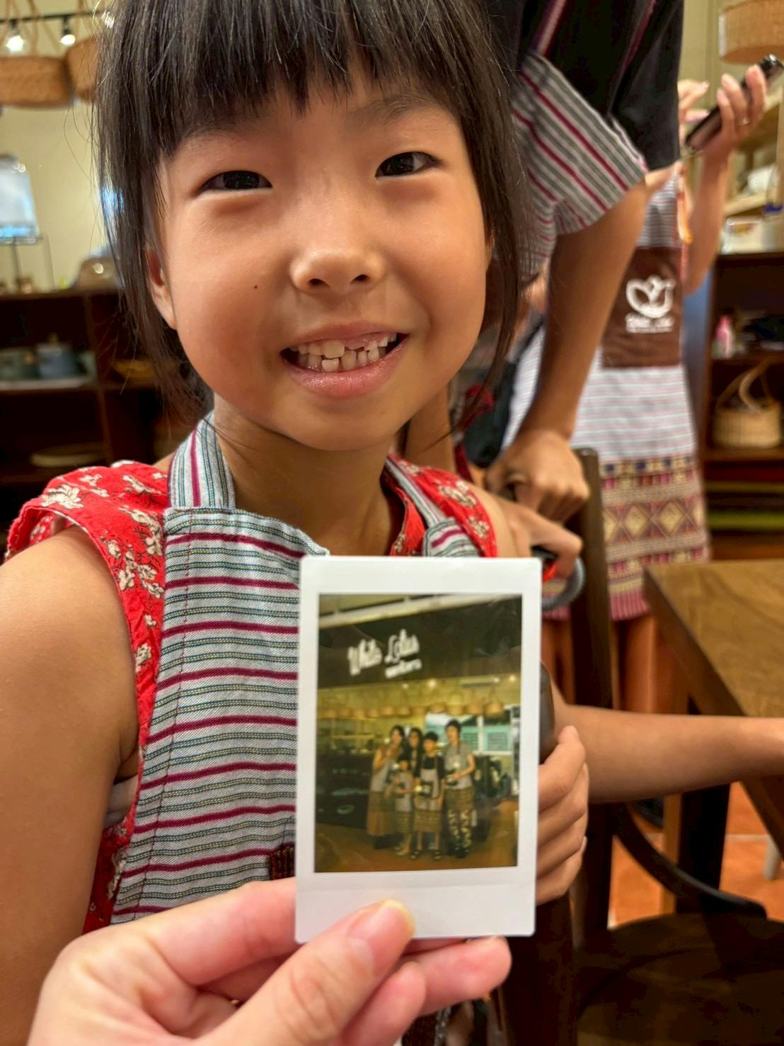 A child smiling while holding a photo in a casual indoor setting with shelves and items in the background.